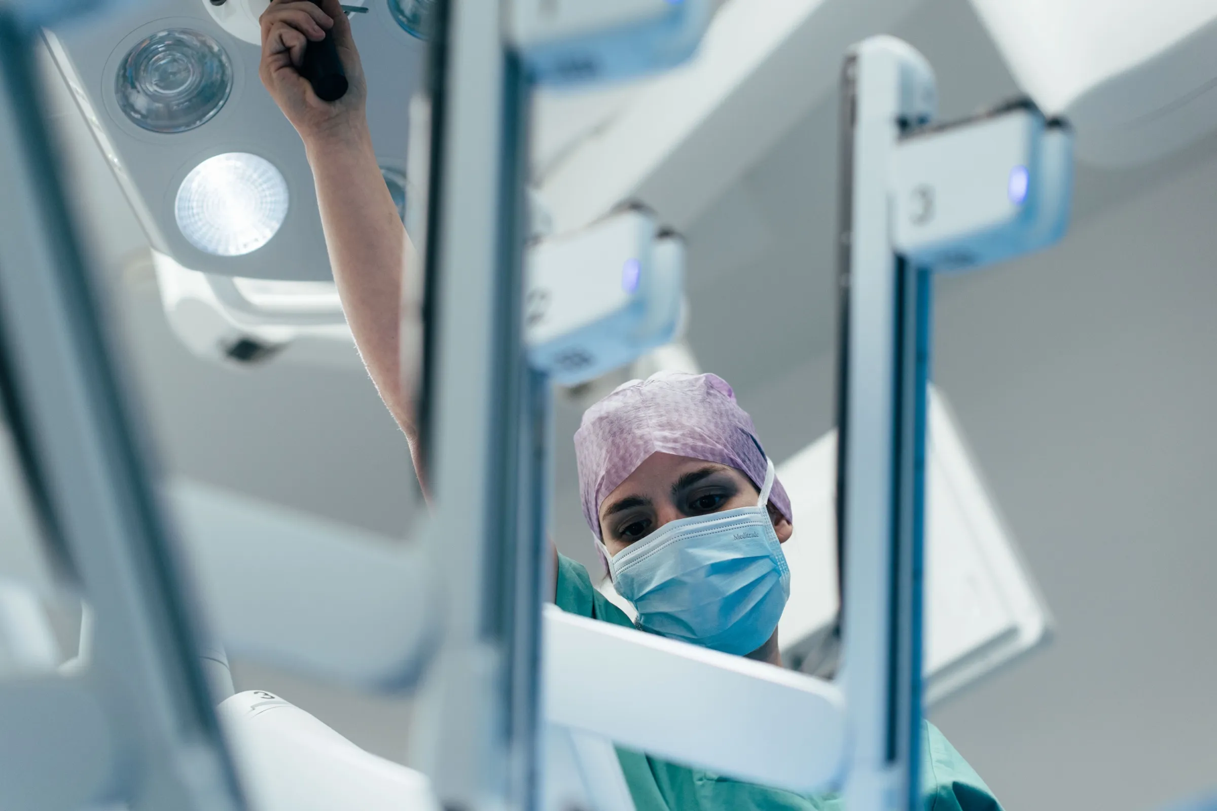 Mitarbeiterin des OP-Teams in Haube und Mundschutz blickt zwischen den Instrumentenarmen des da Vinci Systems hindurch in die Kamera.